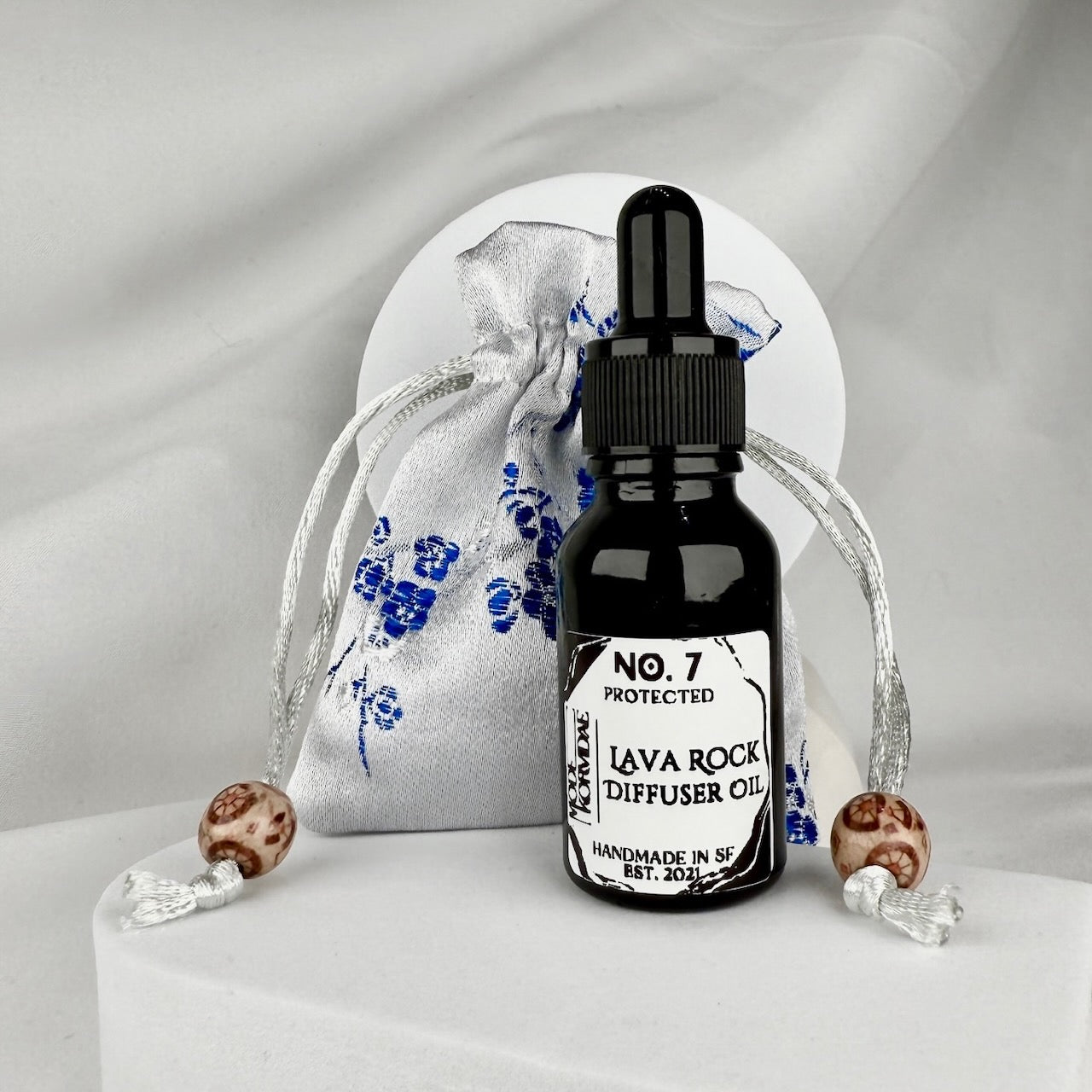 A black glass bottle sitting in front of a white and blue silk sachet pouch on a white background
