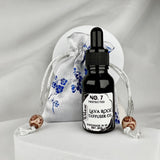 A black glass bottle sitting in front of a white and blue silk sachet pouch on a white background